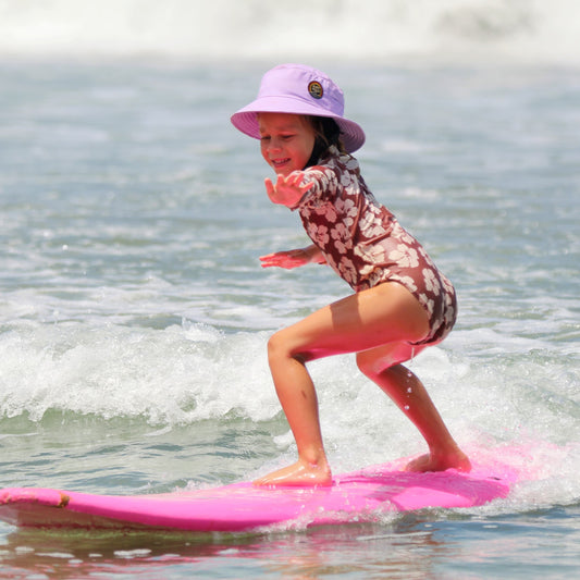 Little surfer girl wearing a lilac bucket hat helmet surfing on a pink surfboard
