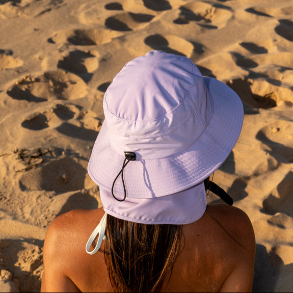 person wearing a lilac pink surf hat with an adjustable back toggle and a removable neck flap seating on the sand