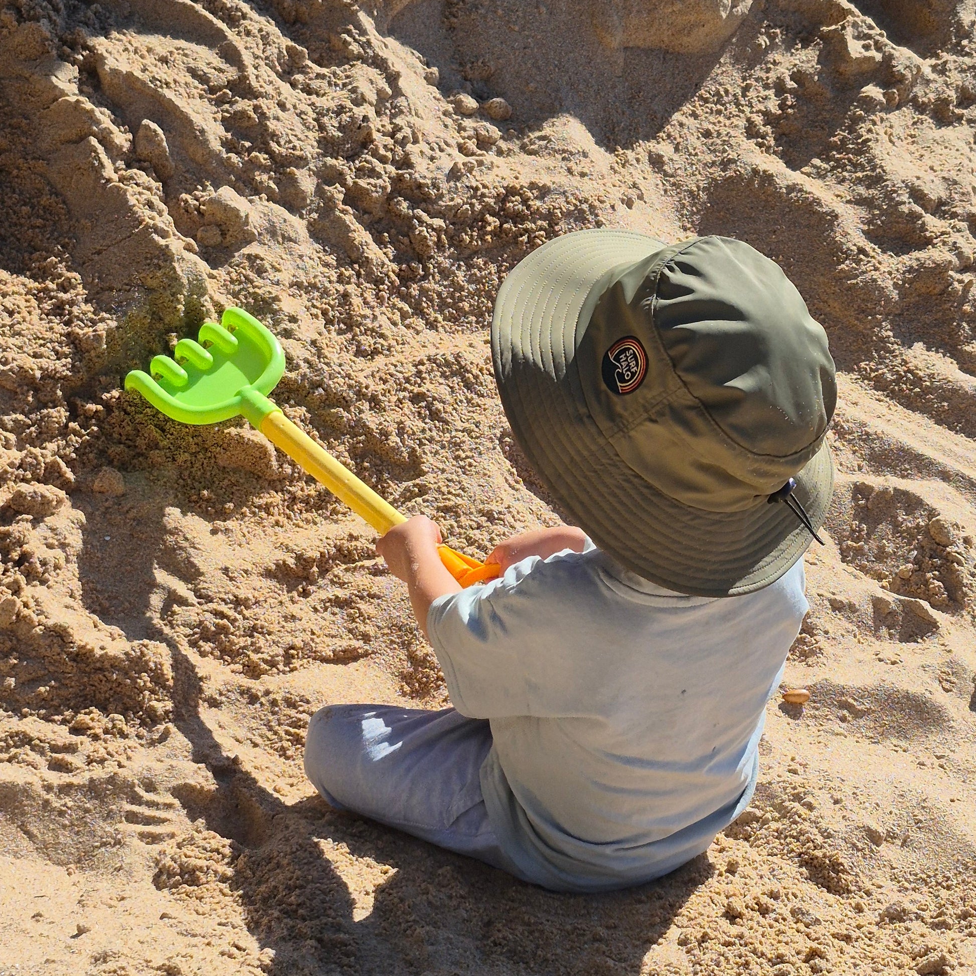 kid playing with sand shovel wearing an olive green surf hat 