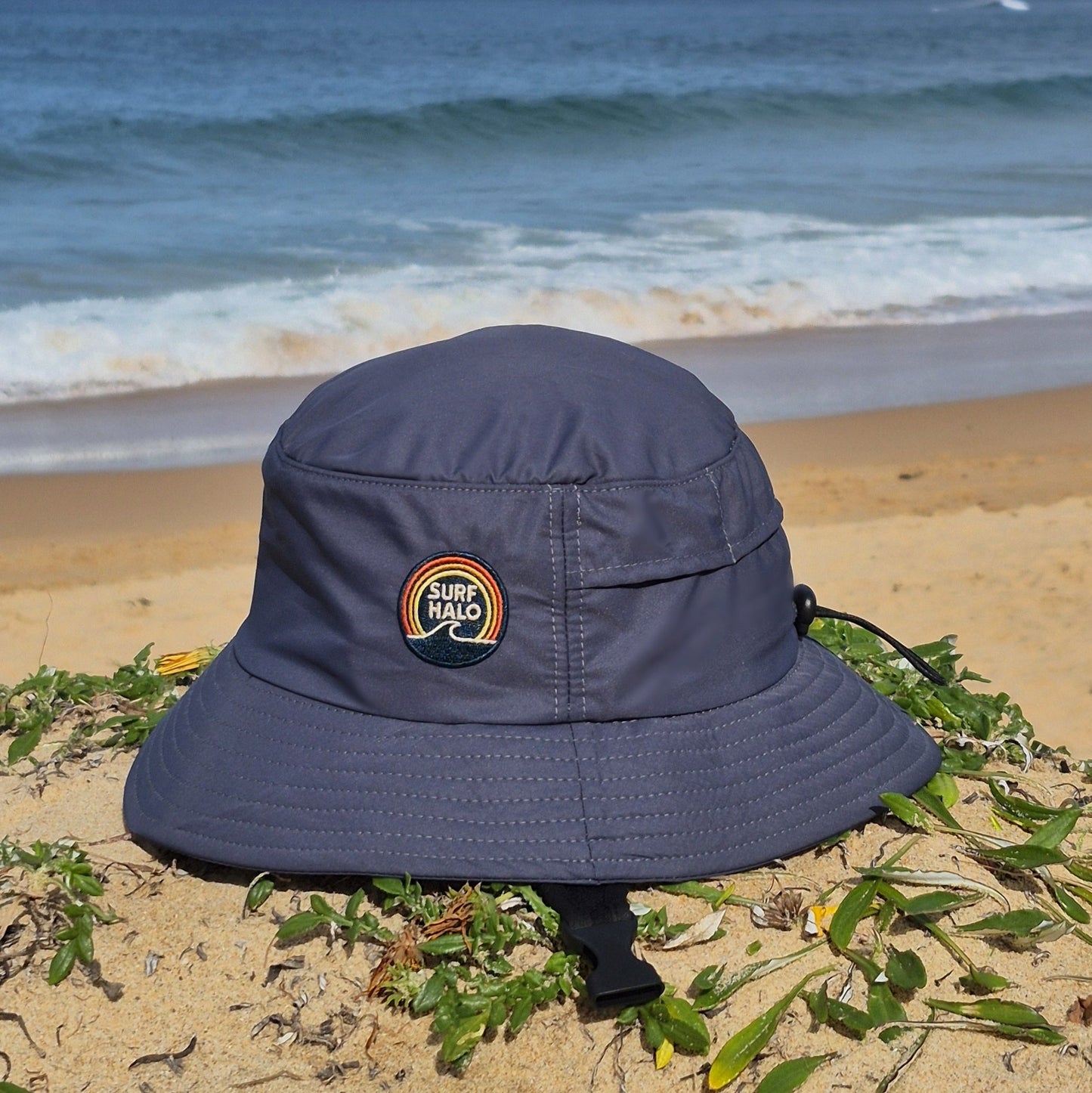 Surf hat helmet charcoal bucket style wide brim UPF5+ adjustable back strap and chin strap in the beach side view