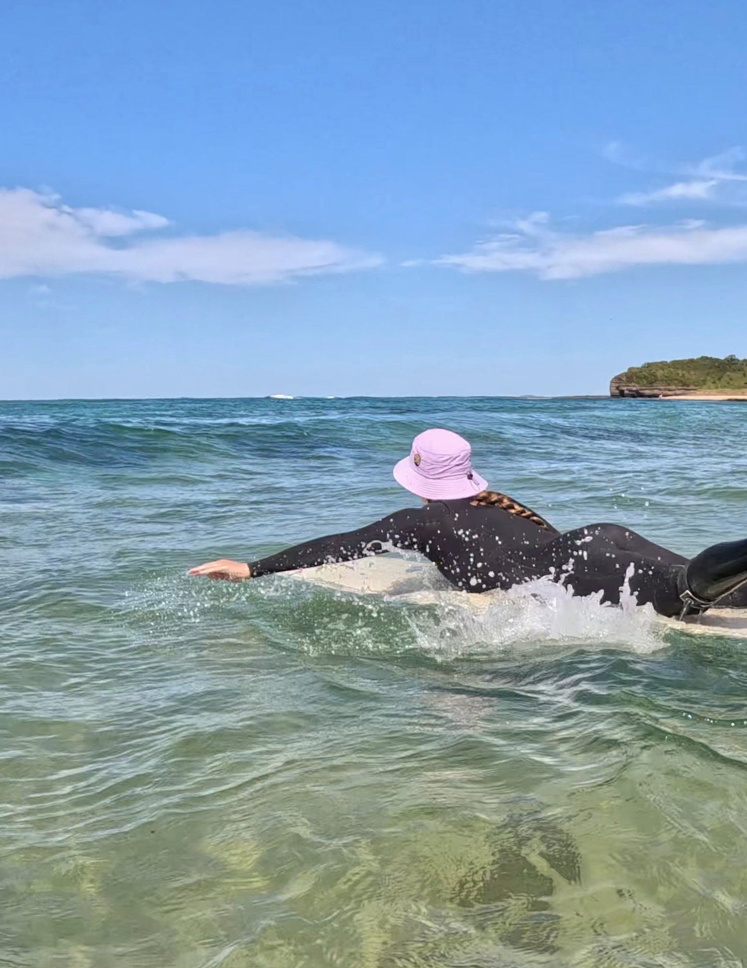 Person lying on a surfboard in clear ocean water with a pink hat