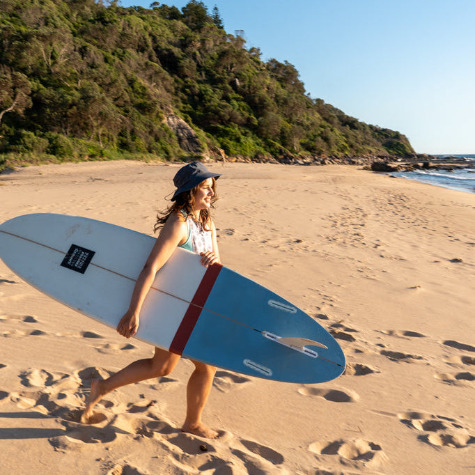 surfer women running to the waves holding a surf board wearing surf hat grey