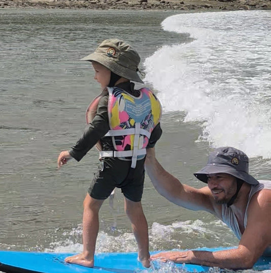 Toddler learning how to surf wearing a surf hat at the beach with his dad