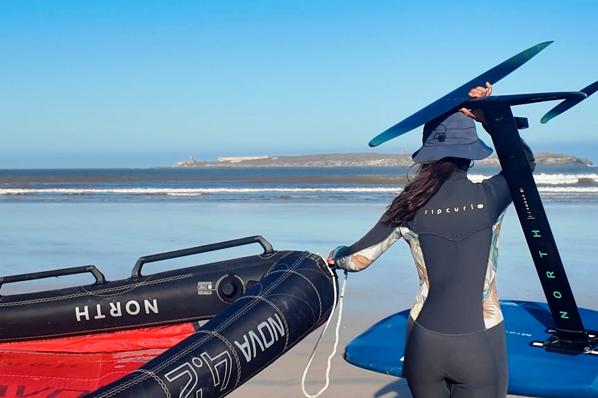 A woman in a full-body wetsuit walks on a sandy beach carrying a black inflatable wing foil tube and handling a blue wing foil setup under a clear blue sky.