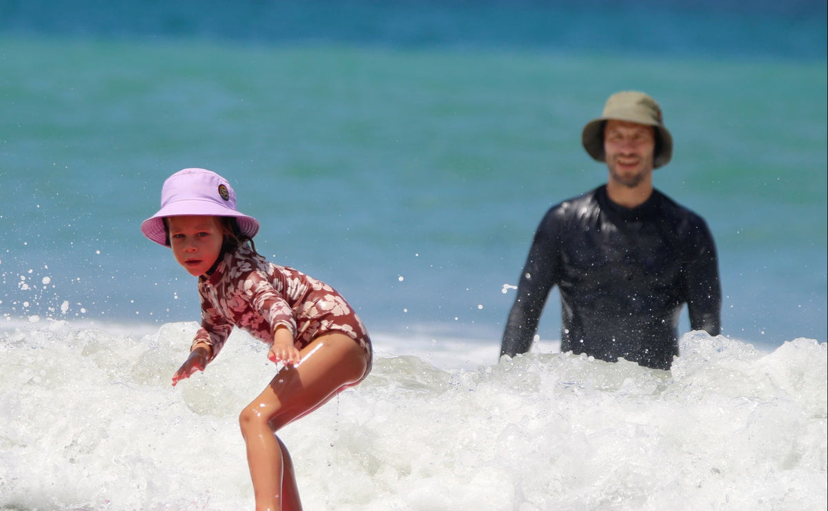 young girl learning how to surf with her dad wearing a surf hat helmet lilac on a pink surfboard at the beach 